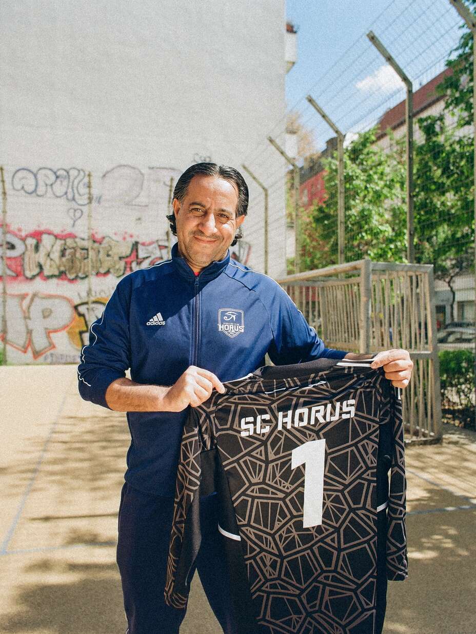 Ein Mann im SC Horus Trainingsanzug hält stolz ein Trikot mit der Nummer 1 vor einem mit Graffiti bedeckten Gebäude hoch.