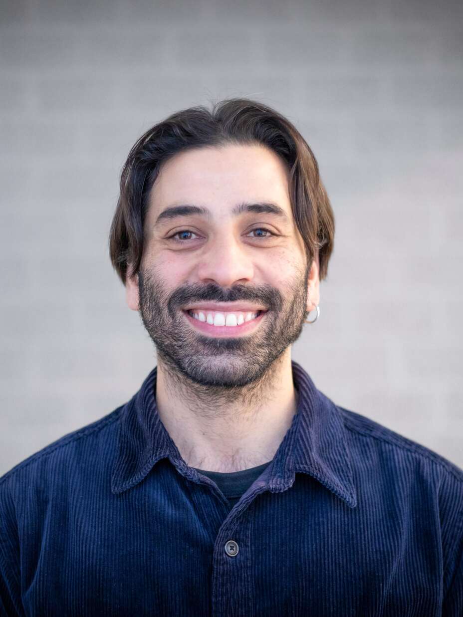 Ein Mann mit Bart lächelt und schaut direkt in die Kamera, vor einem neutralen grauen Hintergrund.