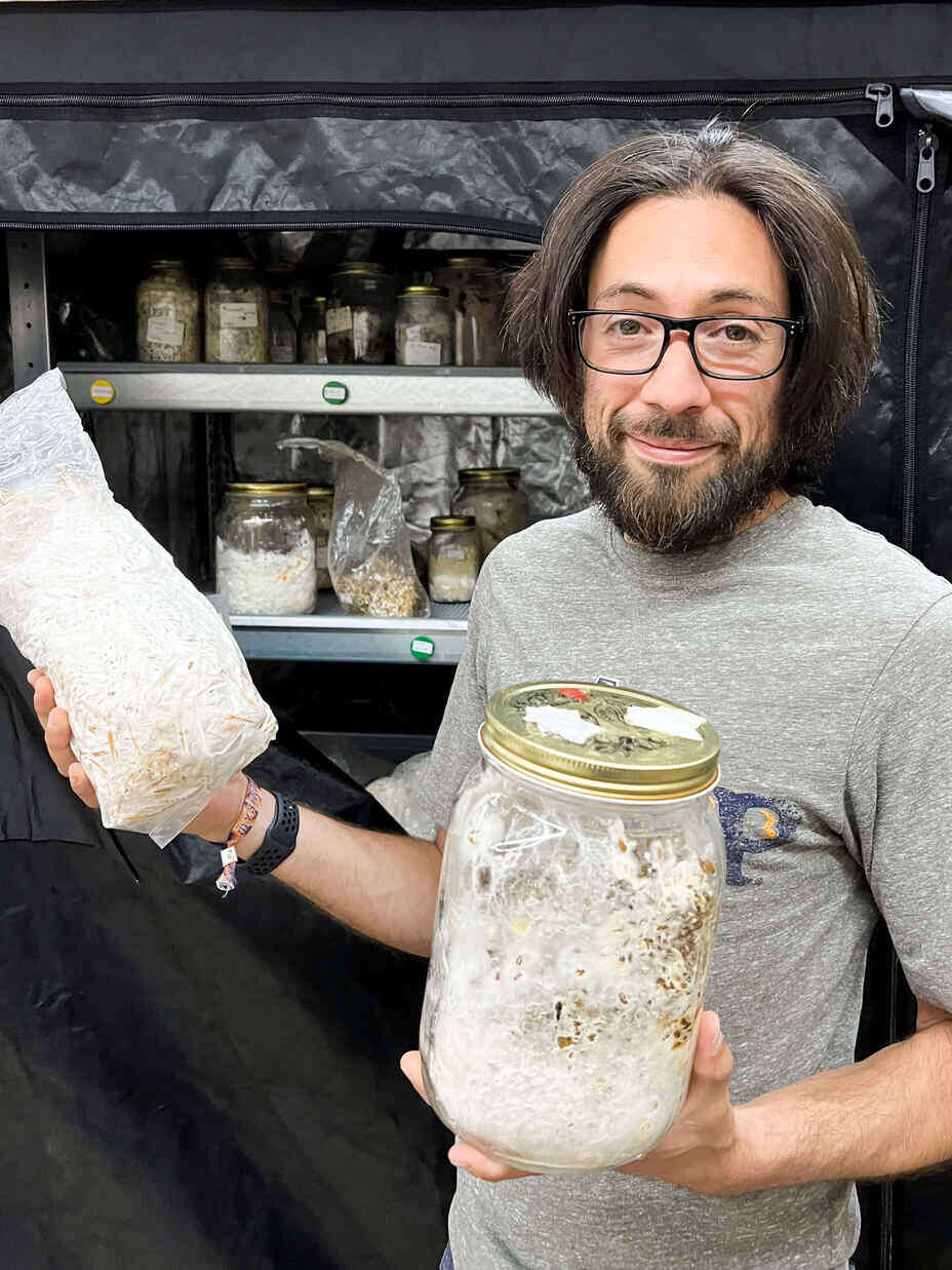 Mann mit Brille und langen Haaren hält Gläser mit Pilzkulturen in einem Lagerraum.