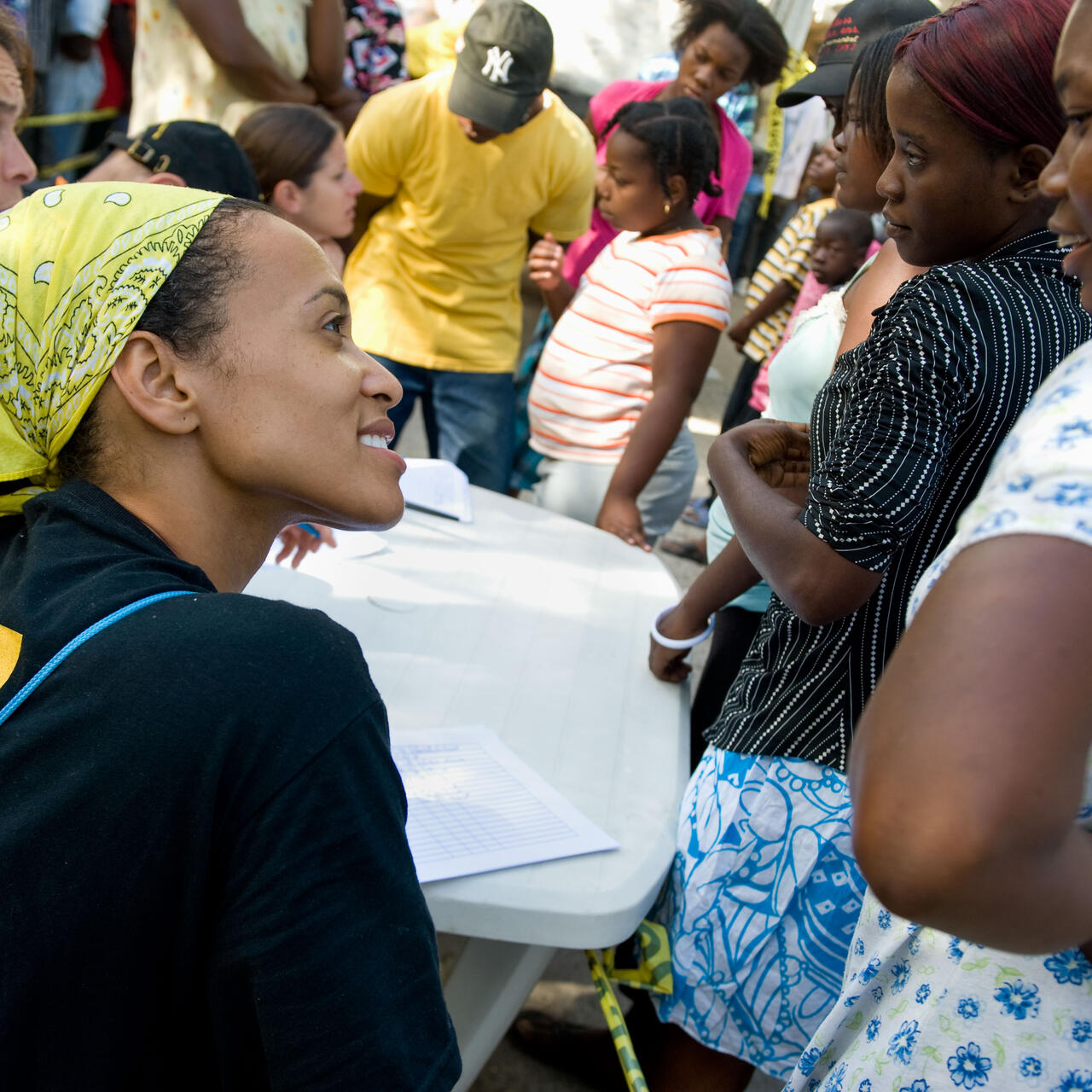 A woman wearing an IRC shirts smiles while interacting with a group of people waiting for humanitarian support.