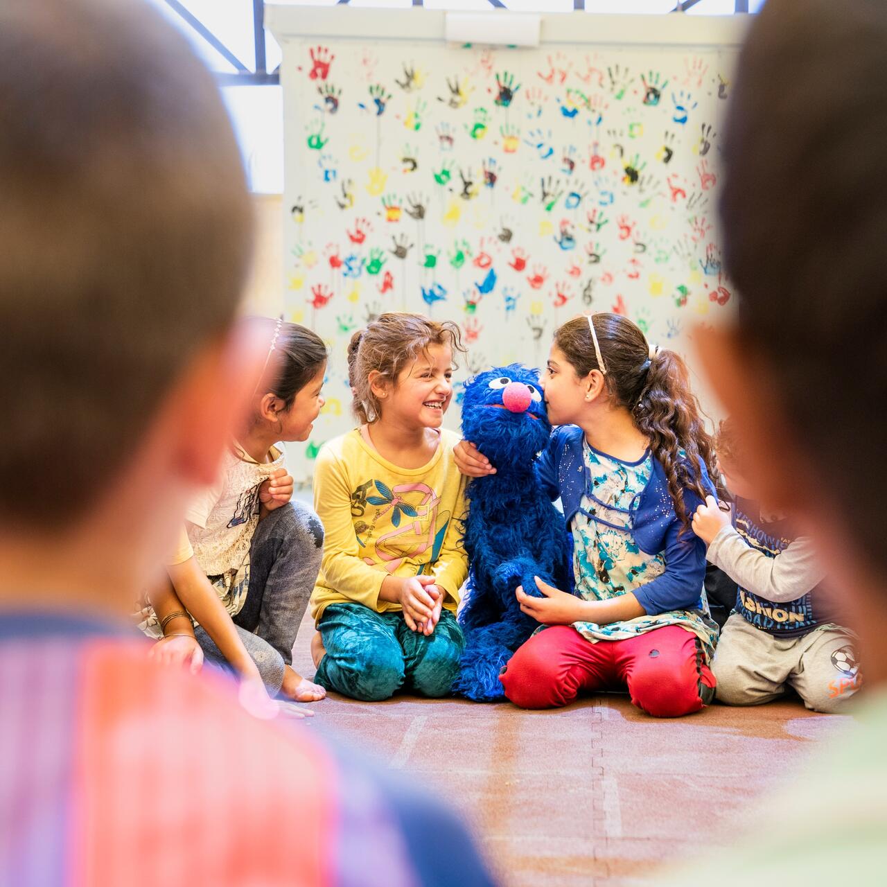 Refugee children sit on the ground in a circle playing witha puppet of the Sesame Street character Grover, a blue monster.