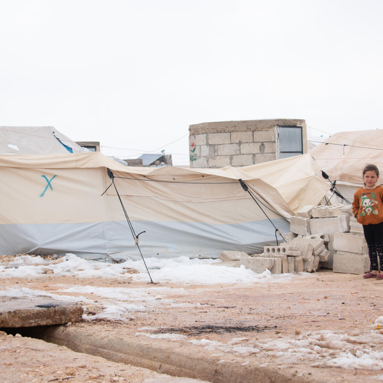 Young girl stands in front of her home in displacement camp, Syria.