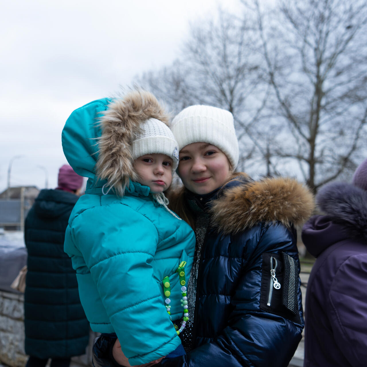 Young girl stands, carrying her baby sister, both facing the camera.