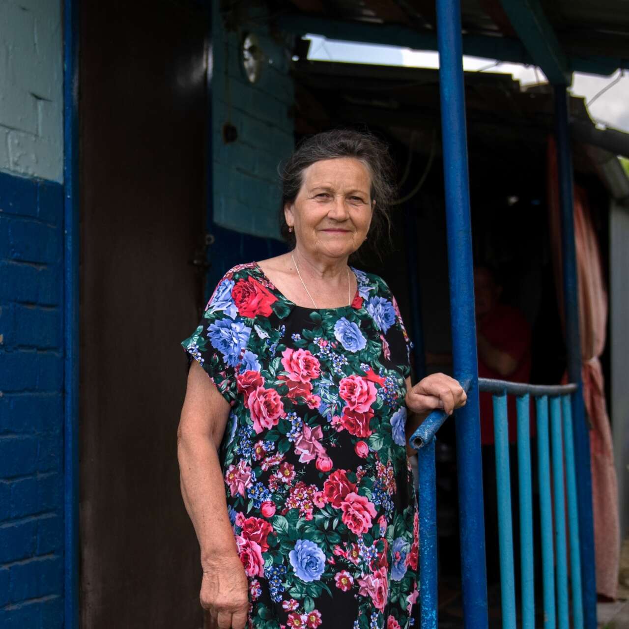 An elderly Ukrainain woman poses for a photo while standing in front of her home.