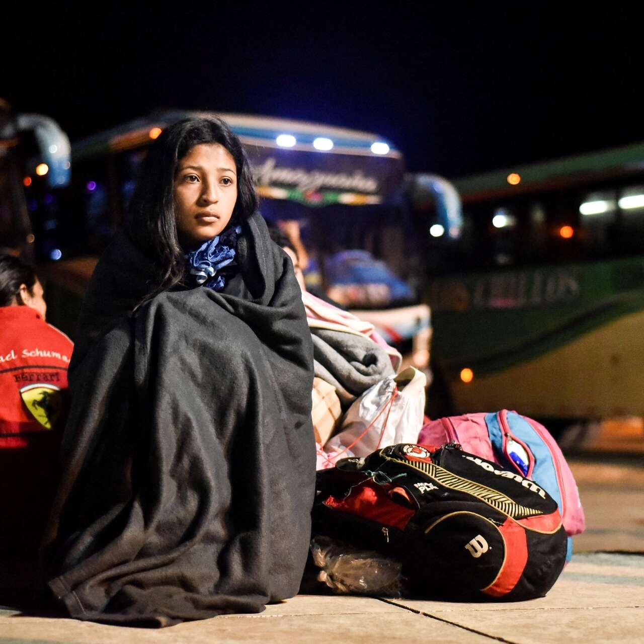 A young woman sits at a bus depot, wrapped in a blanket, next to several bags.