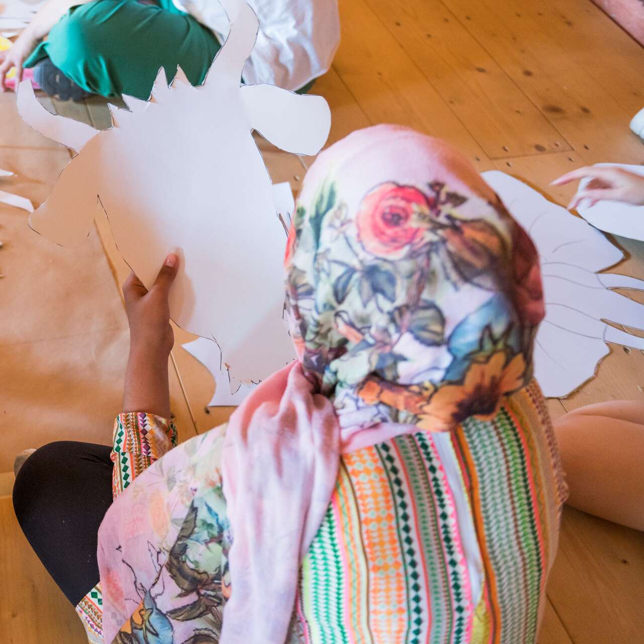 Rückansicht von drei Menschen, die an einem Kunstprojekt auf dem Boden arbeiten; eine Person trägt ein gelbes T-Shirt mit der Aufschrift 'Rescue Aid'.