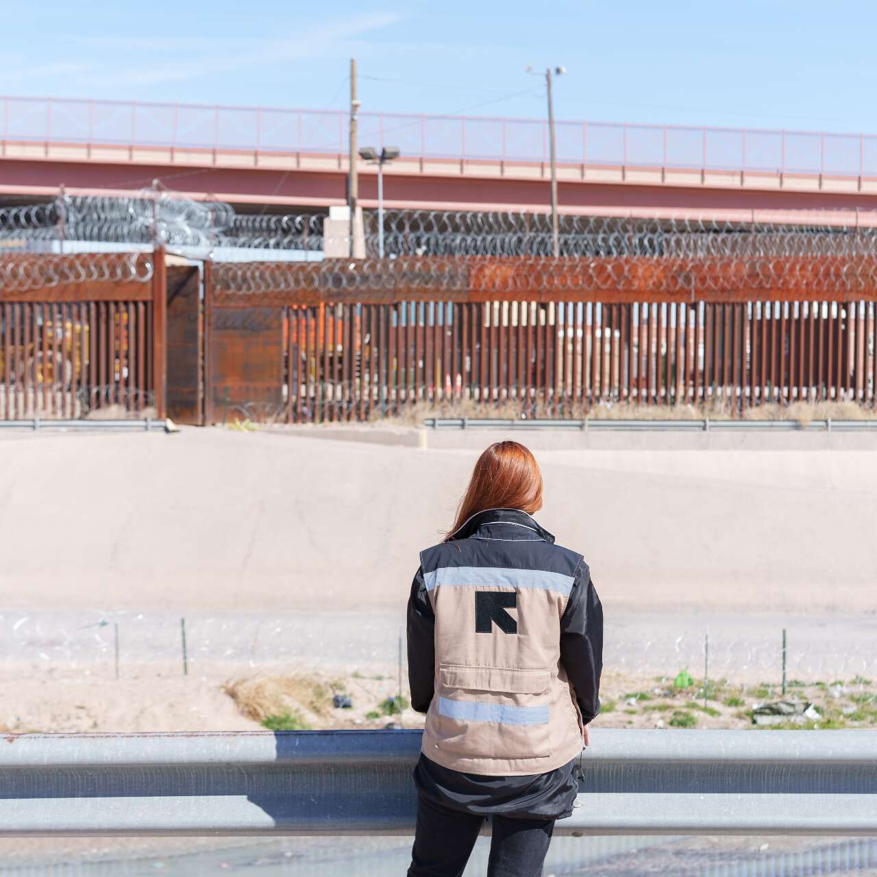 An IRC staff member faces away from the camera and towards the Mexico - U.S. border, while adorning a vest that has the IRC logo on the back.