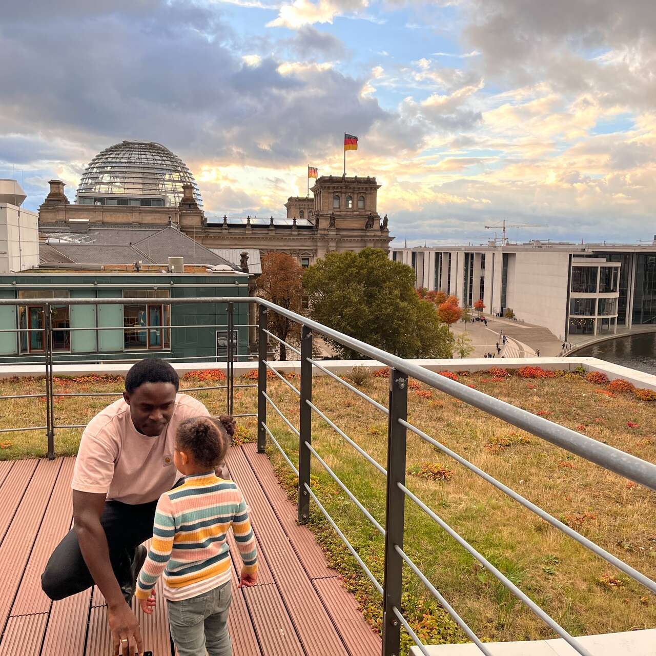 Ein Erwachsener und ein Kind stehen auf einer Terrasse mit Blick auf moderne Gebäude und einen bewölkten Himmel.