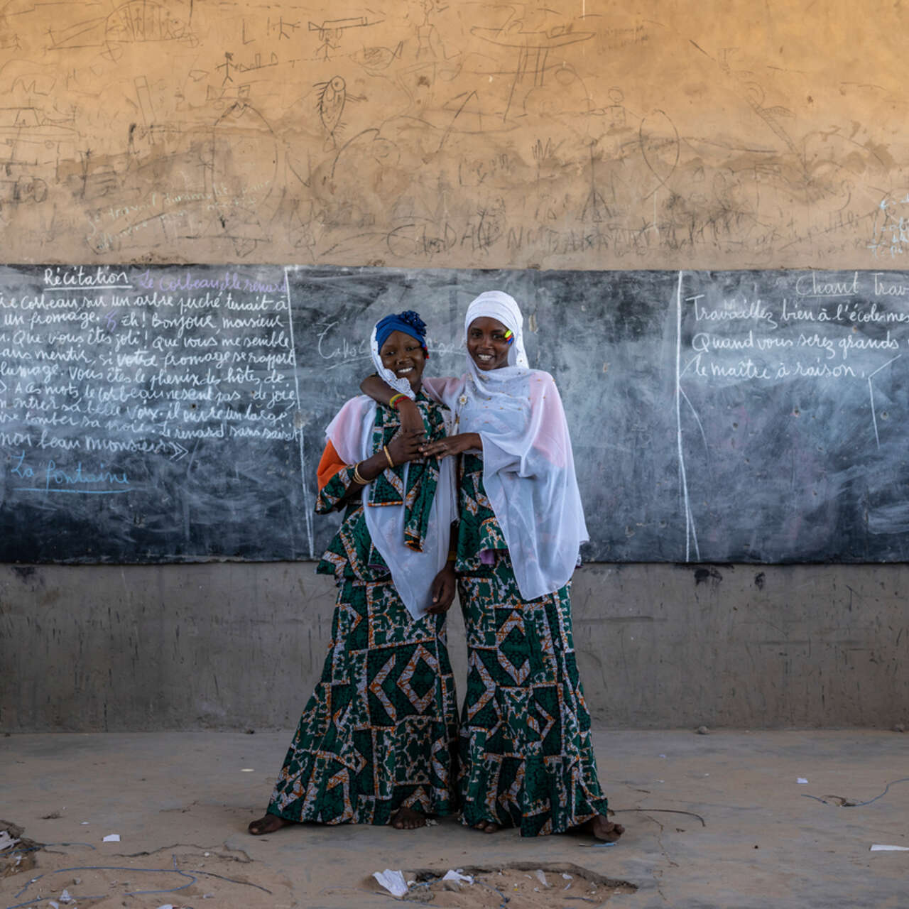 Två tjejer håller om varandra framför en tavla i ett klassrum i ett flyktingläger i Tchad, där RESCUE ger stöd till tonårsflickor.