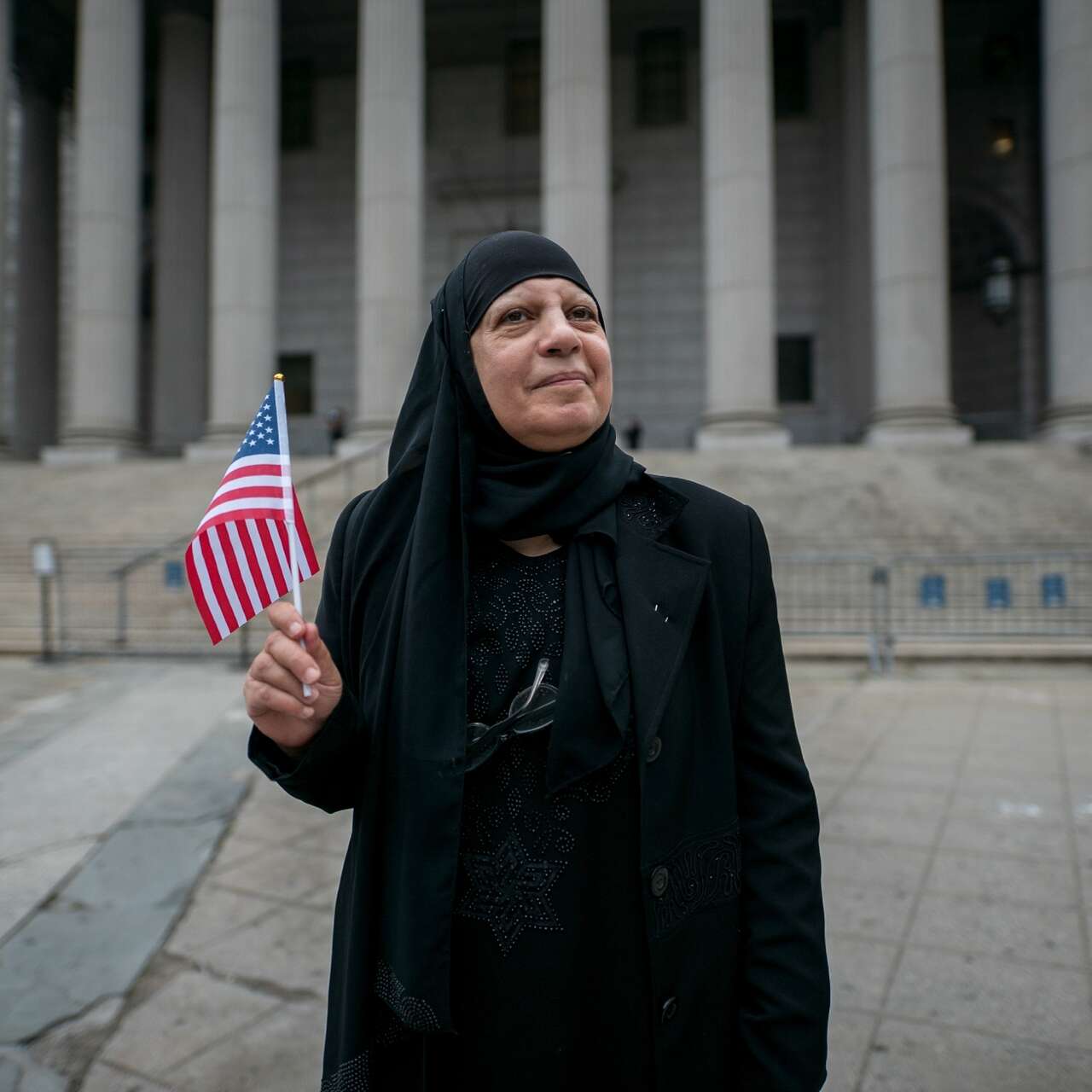 Maha stands outside a Federal office building waving a US flag and smiling.