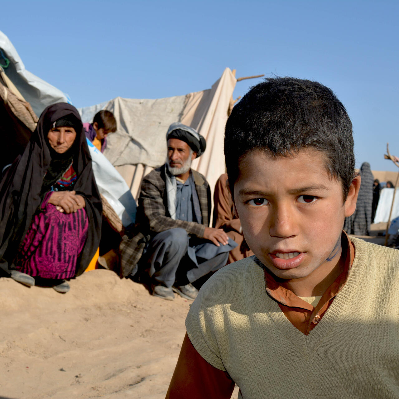 A boy leans toward the camera as an elderly man and woman sit on the sand in front of their tent in a camp for displaced families in Afghanistan.