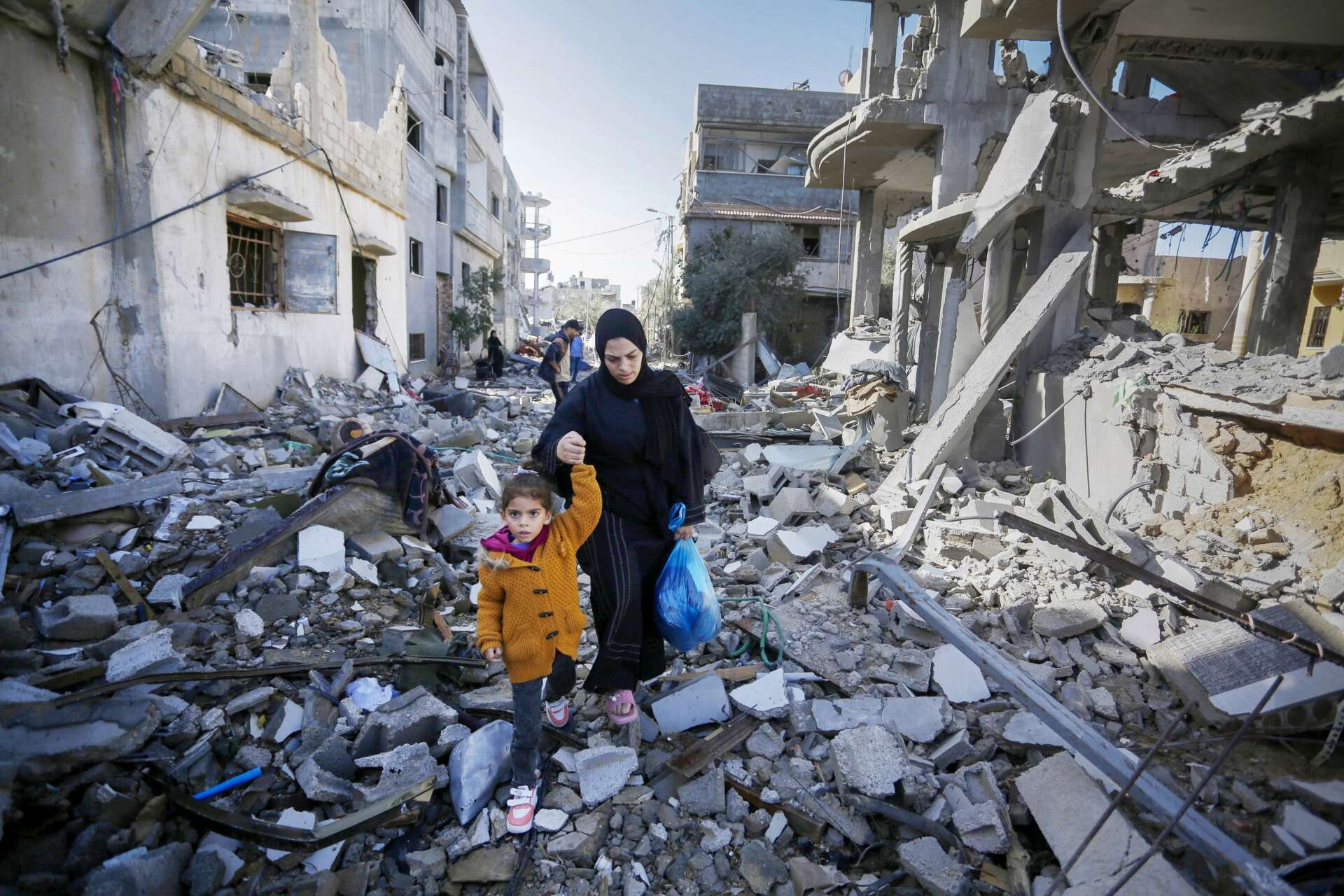 노란 재킷을 입은 어린 소녀와 그녀의 어머니는 전쟁으로 파괴된 가자 지구의 잔재를 걷고 있습니다.