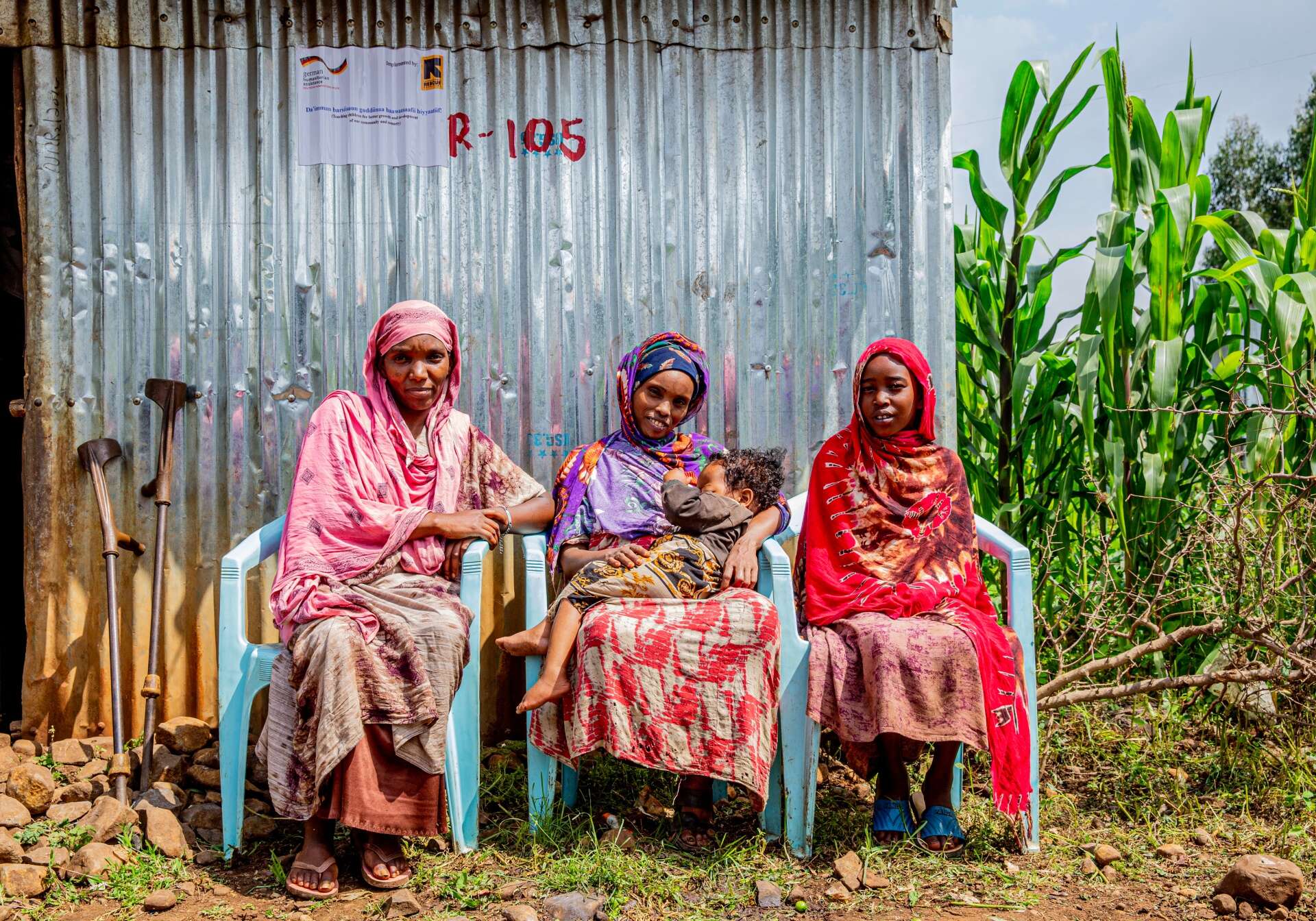 Three women sit in chairs outside and pose for a photo. The woman in the middle holds a young child in her arms.