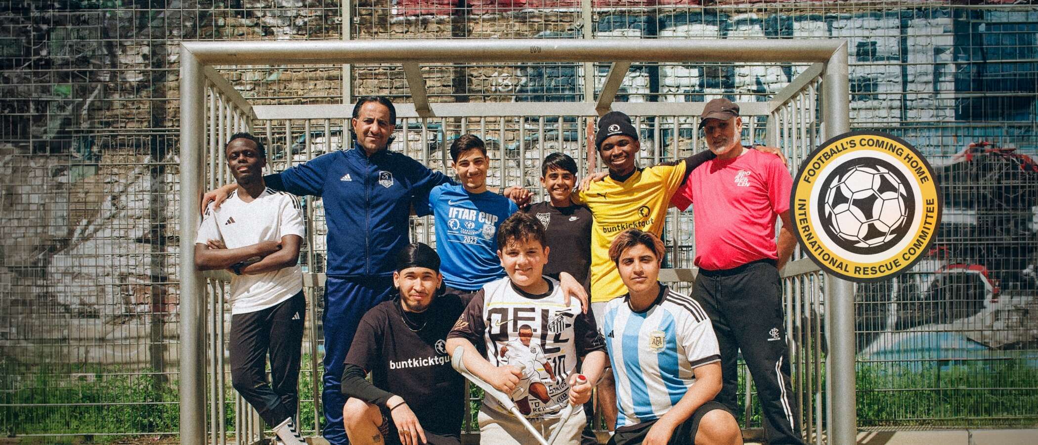 Eine Gruppe von neun Fußballspielern und ihren Trainern steht vor einem Tor auf einem Fußballplatz. Sie lächeln und posieren für das Foto. Rechts unten ist ein Logo mit der Aufschrift 'Football's Coming Home International Rescue Committee' zu sehen.