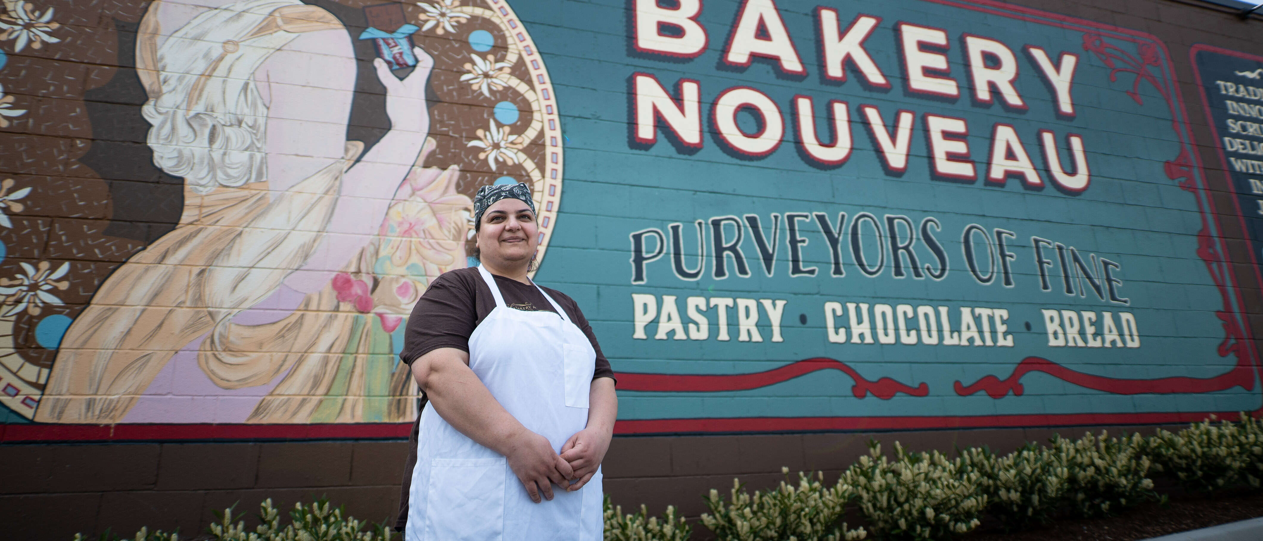 A woman stands by a mural on the side of a bakery and poses for a photo.