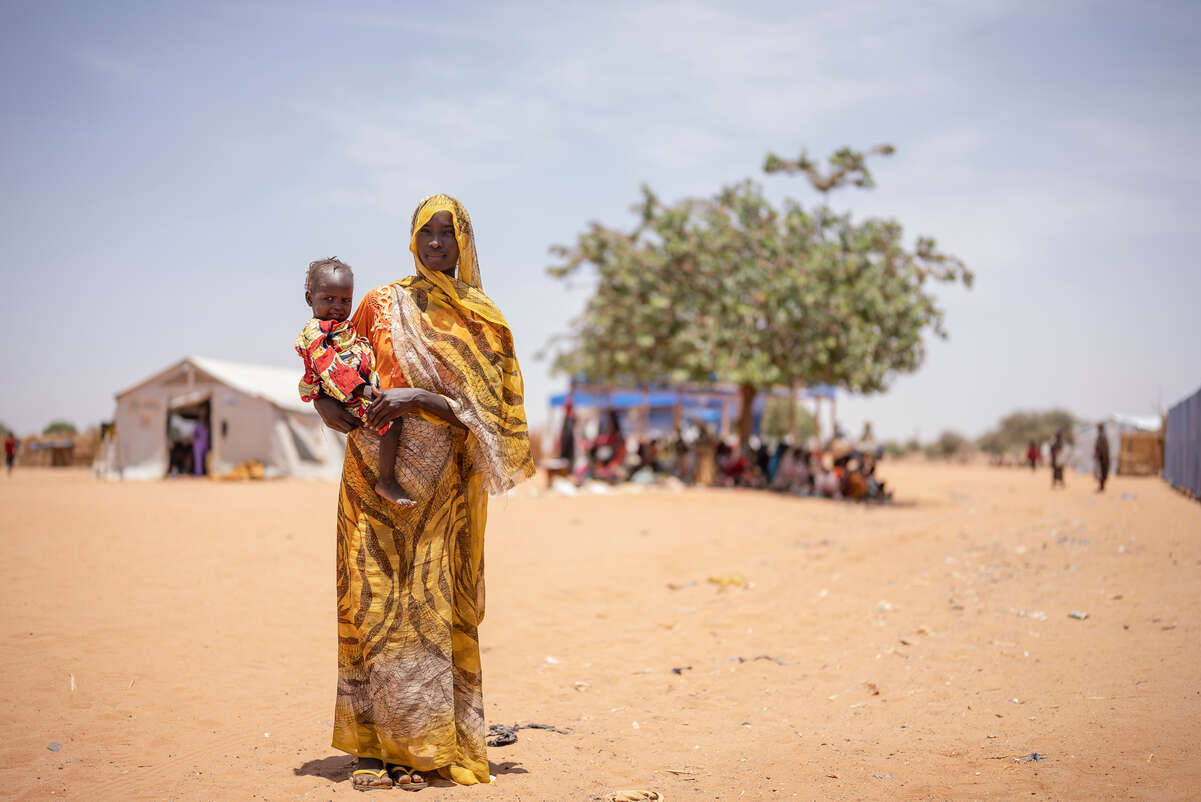 En kvinna på flykt står i ett torrt landskap i Chad och håller sitt barn i famnen. 