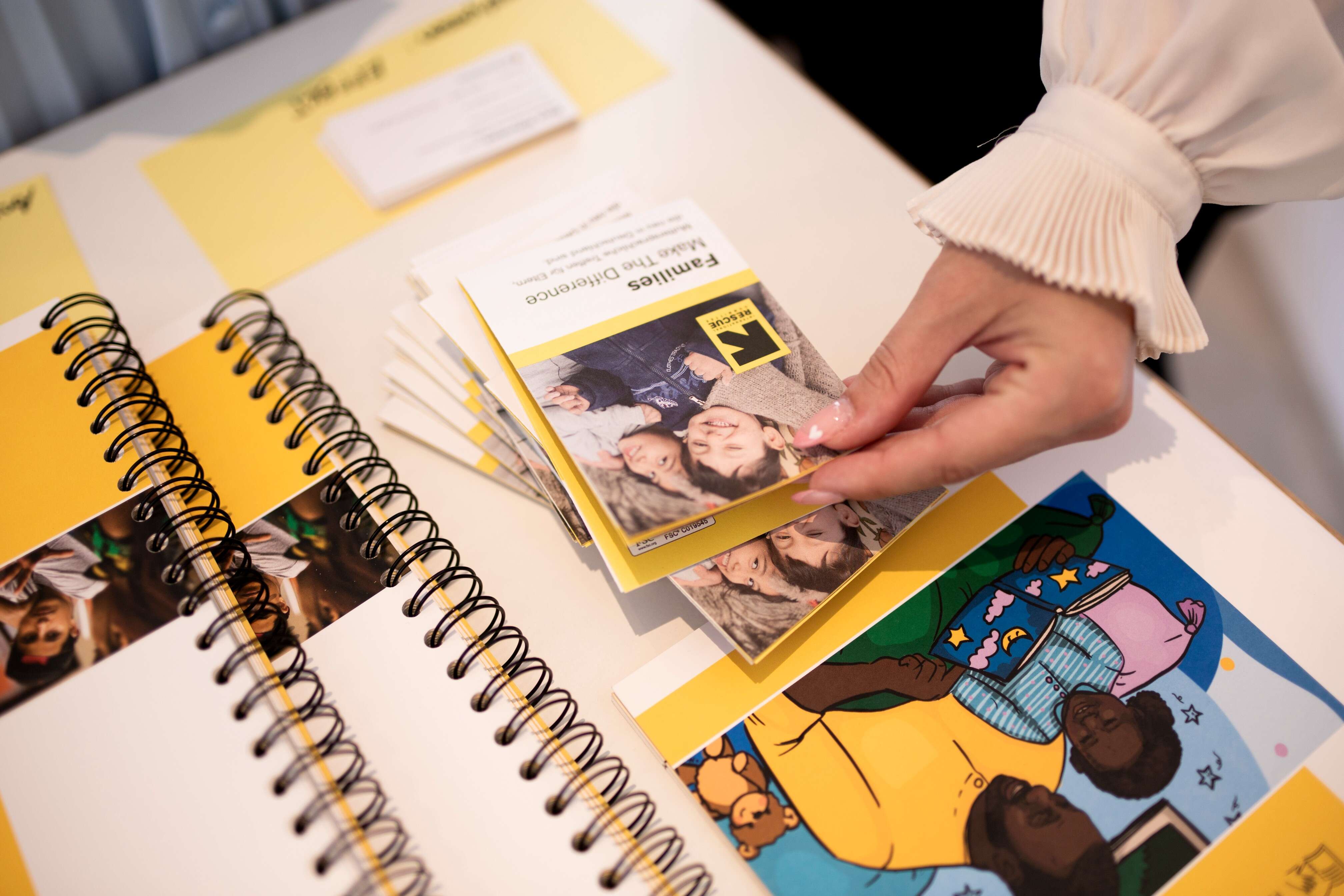 Eine Hand hält ein Faltblatt mit dem Titel "Families Make the Difference" neben einem Tisch mit spiralgebundenen Notizbüchern und bunten Illustrationen.
