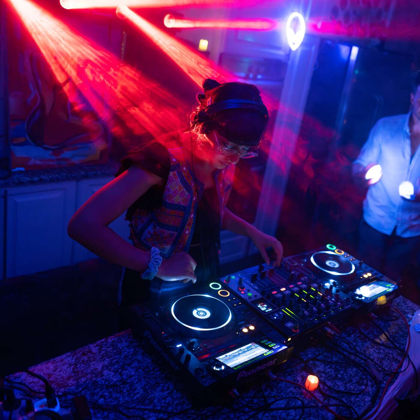 Eine DJane legt in einem Club auf, beleuchtet von bunten Laserlichtern und umgeben von tanzenden Menschen.