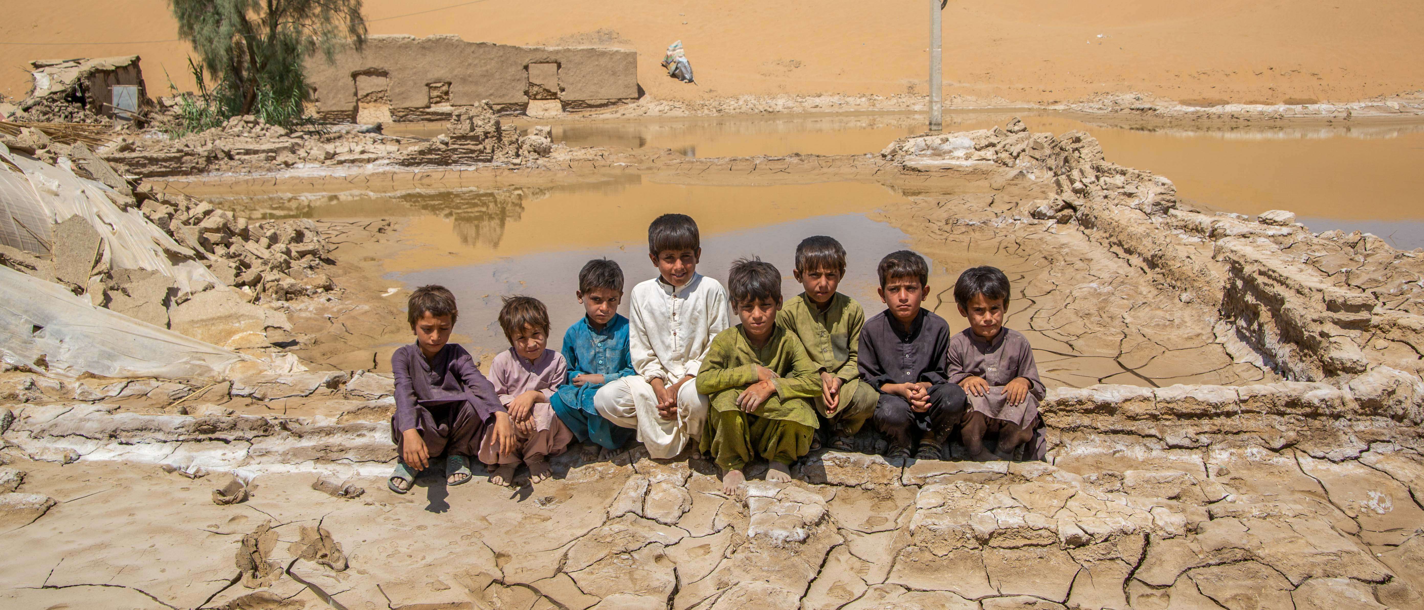 8 boys sit together outside in Pakistan, in front of a landscape devastated by flooding.