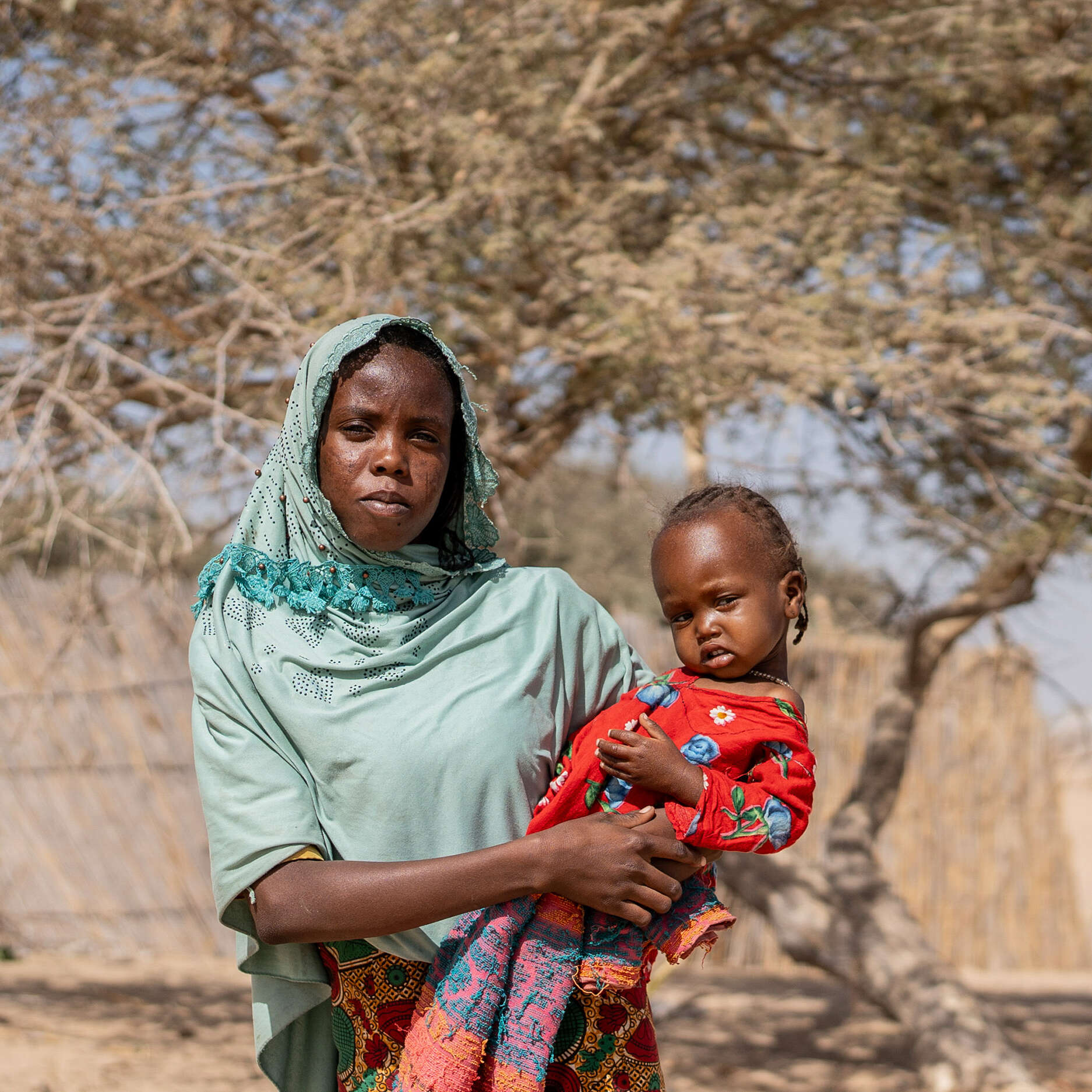 Eine sudanesische Geflüchtete und ihr einjähriges Kind in der trockenen Landschaft vor einer mobilen Gesundheitsstation von IRC in Kafia, Tschad, wo IRC-Mitarbeitende die Tochter auf Unterernährung untersuchten.