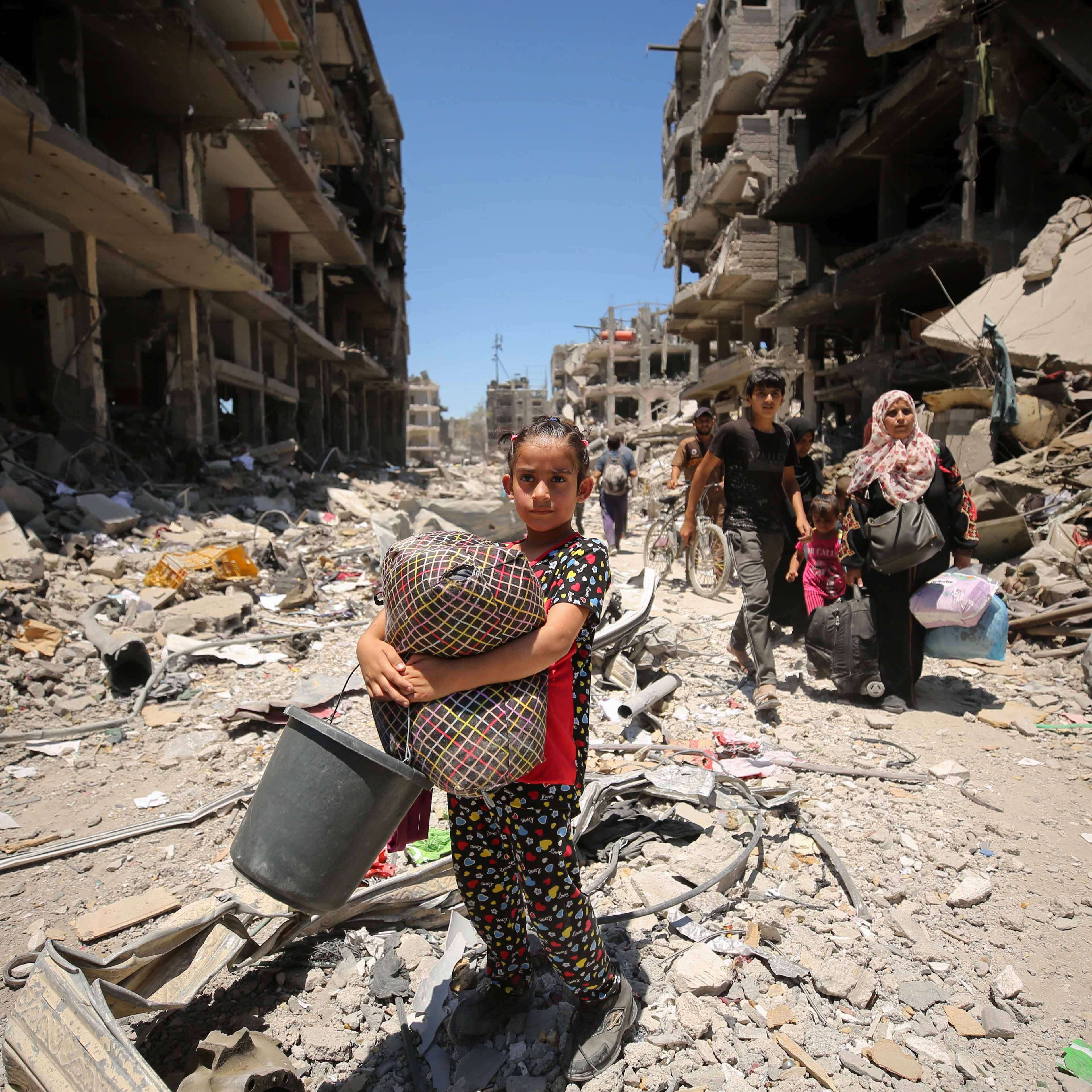 A girl holds belongings as she walks through rubble between destroyed buildings while people follow behind her in Gaza.