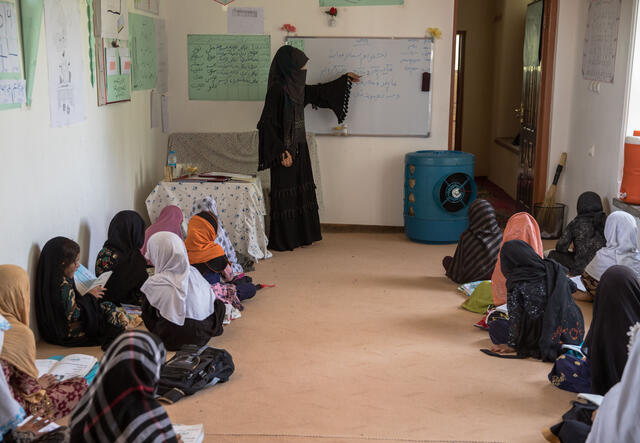 A woman stands at a chalkboard, a group of girls gather and watch the instruction.