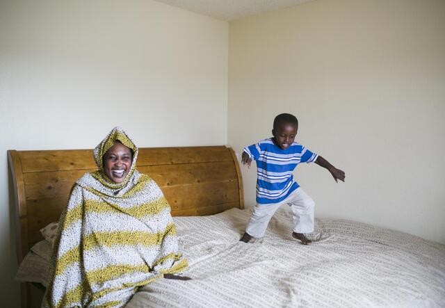 Mom sits on bed laughing and her son runs and jumps across the bed