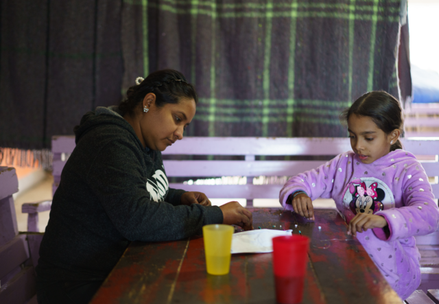 Maria sits across the table from her daughter, watching her make a bracelet.