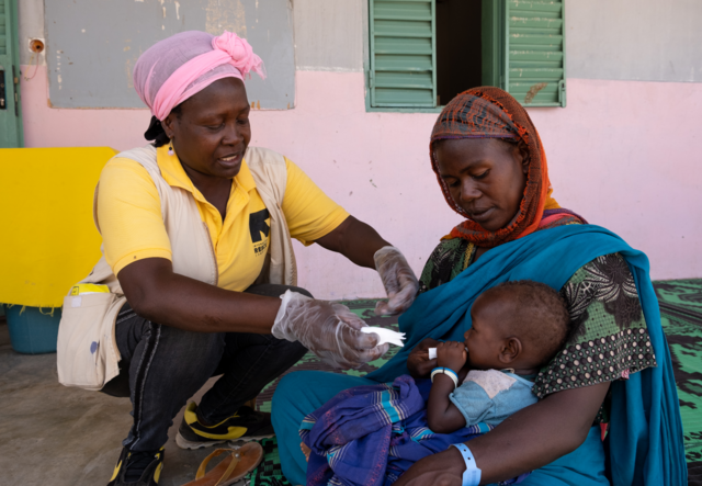 An IRC health worker delivers malnutrition care to a young child who is being held in their mother's arms.