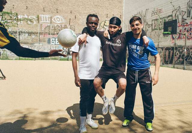 Drei Männer stehen auf einem Fußballplatz, zwei halten sich um die Schultern, während ein vierter Mann einen Ball am linken Bildrand kickt.