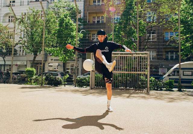 Ein junger Mann, der auf einem städtischen Fußballplatz in sportlicher Kleidung jongliert und dabei einen Fußball in der Luft hält.