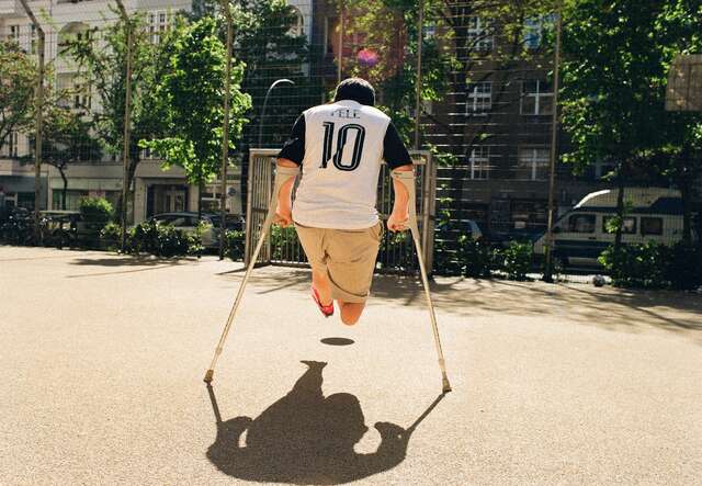 Ein Junge auf Krücken spielt Fußball auf einem sonnigen Spielplatz, trägt ein Fußballtrikot mit der Nummer 10.