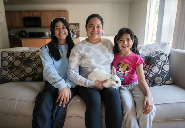 A mother sits on a couch and poses for a photo next to her two daughters. She holds the family's pet bunny in her lap.