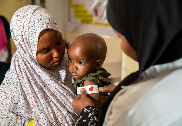 한 어머니가 국제구조위원회 영양담당 직원이 영양실조 징후를 검사하기 위해 자녀를 안고 뮤악테이프로 팔 둘레를 측정하는 것을 돕고 있습니다. 