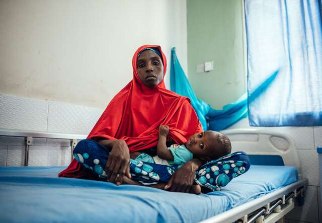 Hasfat hält ihren Sohn im Arm, während sie auf einem Bett in einer medizinischen Einrichtung in Nigeria sitzt.