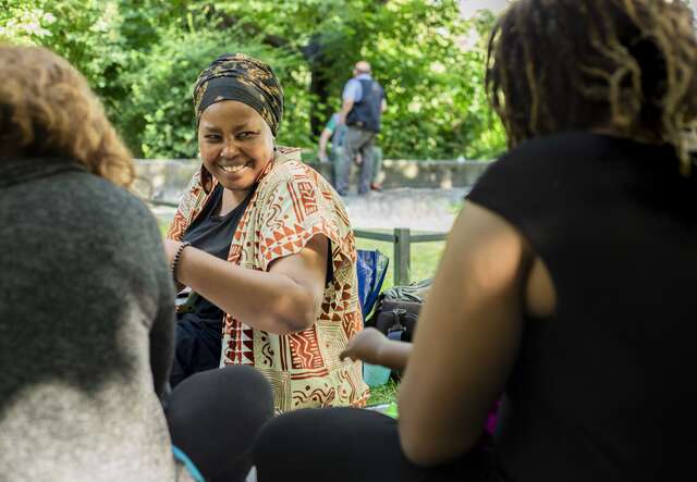 Eine Frau lacht und spricht mit anderen Menschen im Freien, umgeben von grünen Bäumen.
