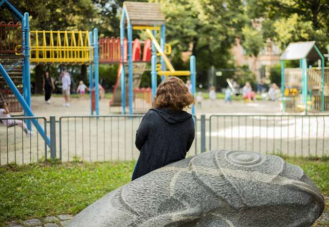 Eine Person sitzt auf einer Bank in einem Park und schaut in Richtung eines Spielplatzes mit bunten Spielgeräten.