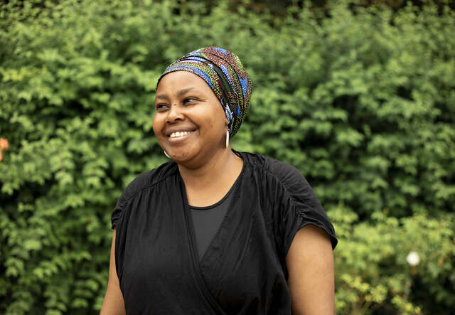 Eine Frau mit einem Stirnband lächelt und steht vor einer grünen Hecke.