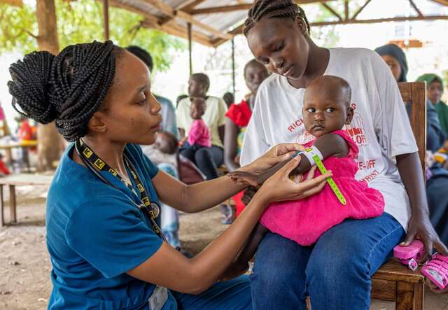 Ein IRC-Gesundheitsarbeiter untersucht ein junges Mädchen auf Anzeichen von Unterernährung in einer Außenklinik in Kenia. Das Kind sitzt auf dem Schoß seiner Mutter, während ein Unterernährungshelfer die Untersuchung durchführt.