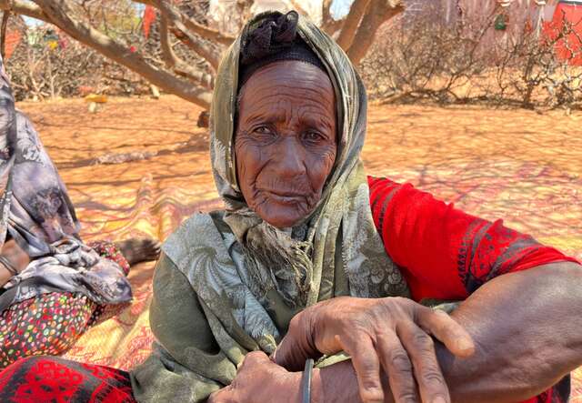 An older woman sits on the ground with her hands crossed and poses for a portait.