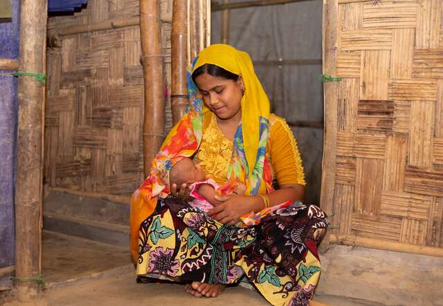 A mother kneels on the floor of her home in Bangladesh and holds her young child in her arms.