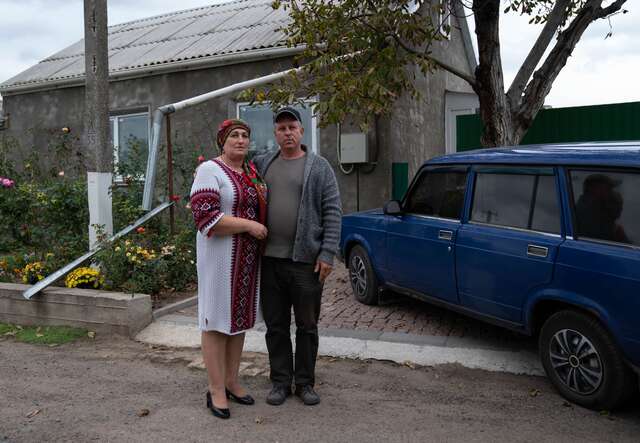 Olga posiert für ein Foto neben ihrem Mann vor ihrem Haus in der Ukraine. Obwohl ihr Haus in gutem Zustand ist, hat der Krieg ihre Gemeinde stark in Mitleidenschaft gezogen und die beiden gezwungen, für längere Zeit unter der Erde Schutz zu suchen.