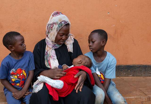 A mother holds her newborn child in her arms. Her two sons sit on either side of her, looking at the newborn child.