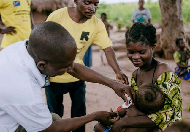 A woman holding her baby in her arms and two IRC workers test the baby for malnutrition