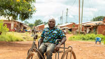 A man poses for a photo while sitting in a wheelchair and hand-cranked bicycle.