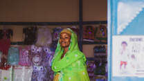 A woman, wearing a vibrant green headscarf and yellow dress, smiles in a shop in Cameroon.