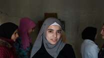 A young woman smiles at the camera in Lebanon. Behind her, other young women speak to an IRC staff member.