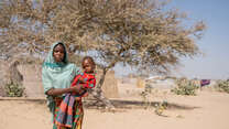 A mother holds her child on her hip in front of a barren back drop.