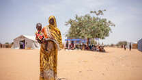 En kvinna på flykt står i ett torrt landskap i Chad och håller sitt barn i famnen. 