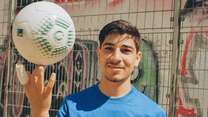 Ein junger Mann in einem blauen „Iftar Cup“-T-Shirt balanciert einen Fußball auf seinem Finger vor einer Wand mit Graffiti.